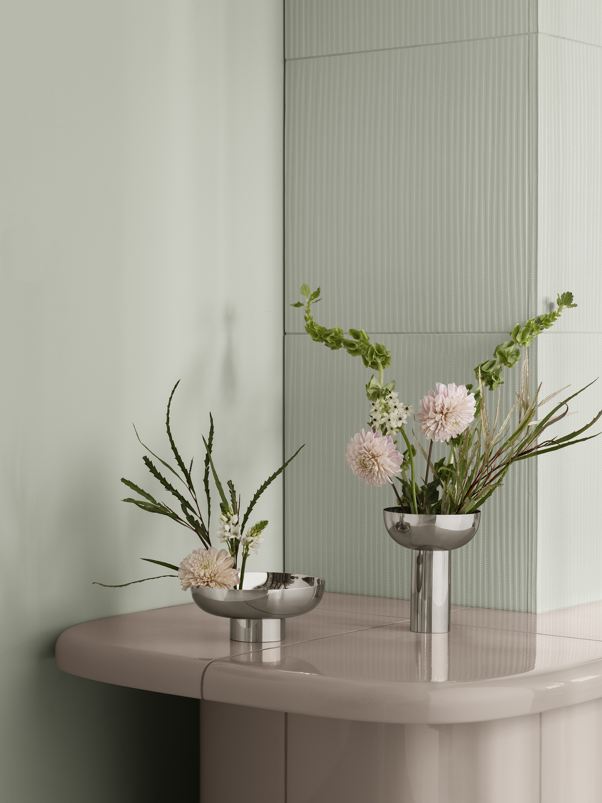 Two modern silver vases with assorted flowers on a glossy beige table against a textured pale green wall. The setting is serene and elegant.