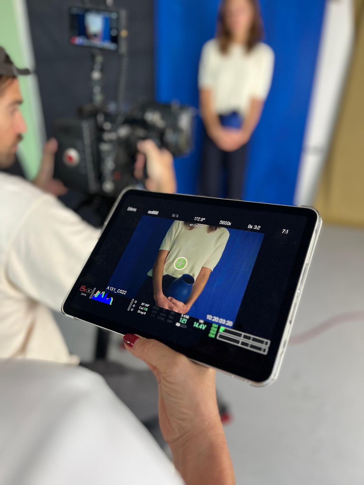 Coulisses d’un shooting produit avec une équipe caméra filmant un modèle tenant un vase bleu, suivi sur une tablette.