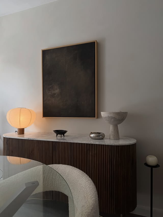 Buffet en bois foncé avec plateau en pierre claire, lampe en papier allumée et coupes décoratives sous un tableau abstrait.