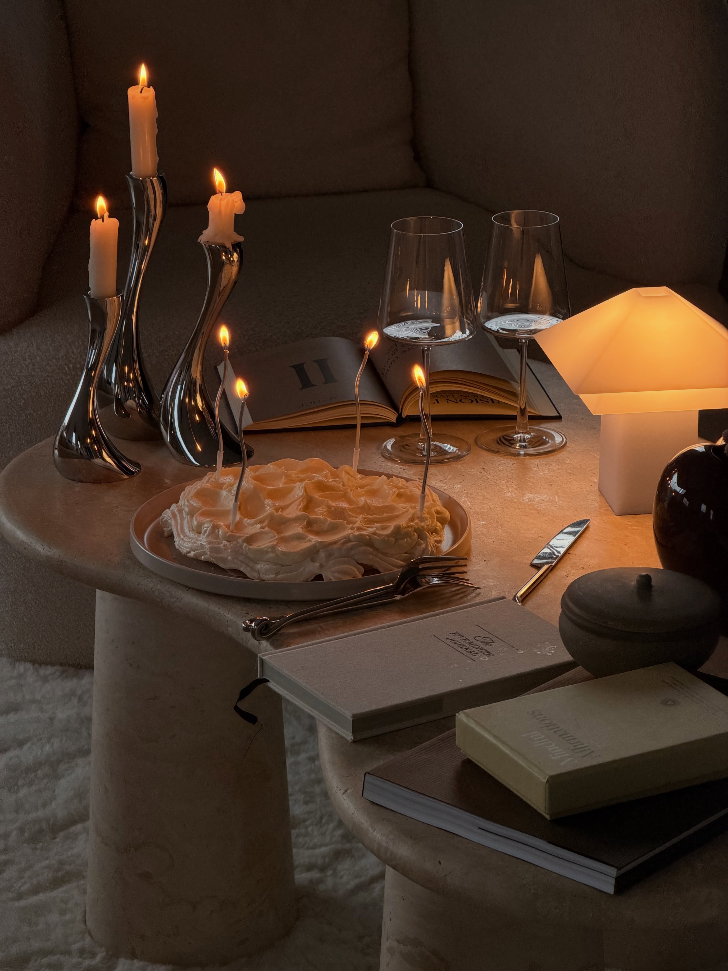 Silberfarbene Kerzenhalter, eine leuchtende Tischlampe und eine Torte mit brennenden Kerzen auf einem beigen Couchtisch.